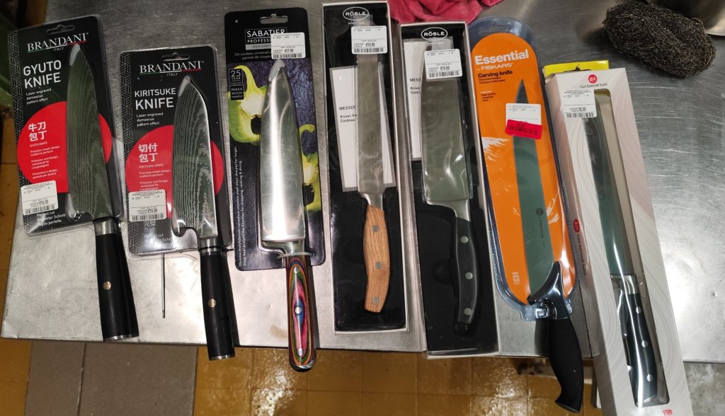 Seven new budget kitchen knives still in their packaging, lined up on a stainless steel restaurant counter: Brandani gyuto and kiritsuke, Sabatier chef's knife, two Rösle knives, a Fiskars carving knife and a Carl Schmidt Sohn bread knife